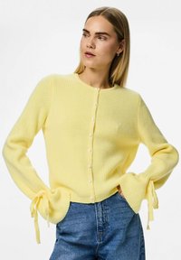 Gul ribbad cardigan med pärlknappar, rund halsringning och utsvängda ärmar som är knutna vid handlederna. Kombinationen med denimjeans förbättrar utseendet.