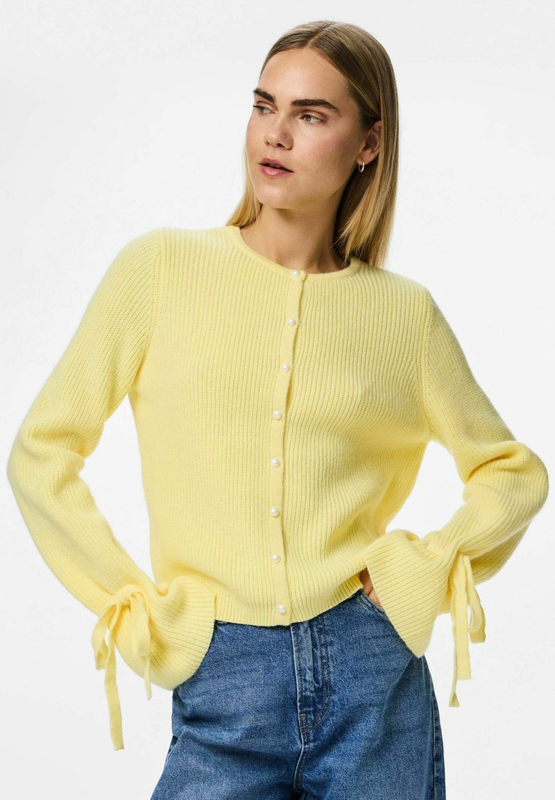 Gul ribbad cardigan med pärlknappar, rund halsringning och utsvängda ärmar som är knutna vid handlederna. Kombinationen med denimjeans förbättrar utseendet.