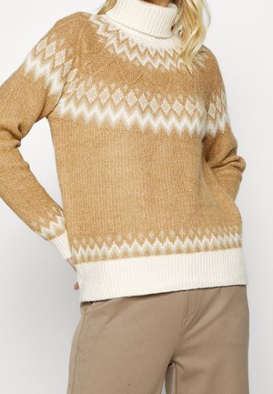 Kvinde iført en beige og hvid mønstret rullekravesweater med zigzag-design og beige bukser, stående med hænderne på hoften.