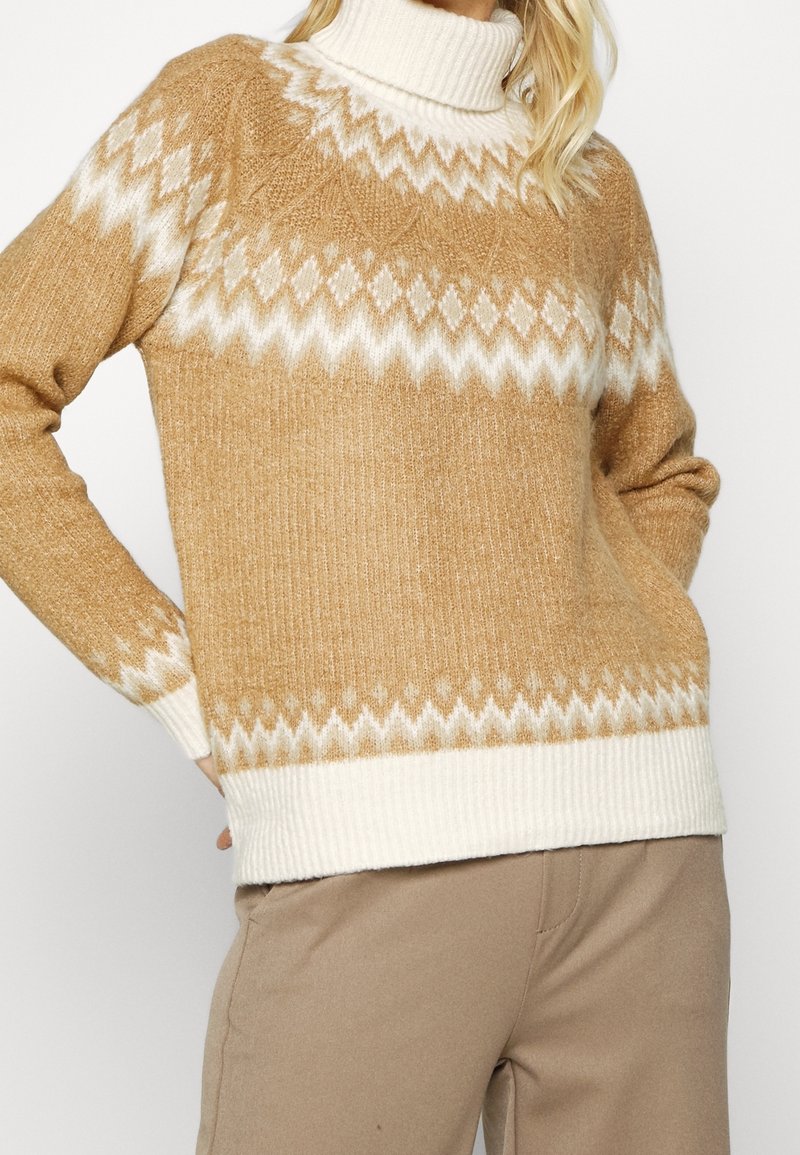 Femme portant un pull à col roulé beige et blanc à motif zigzag et un pantalon beige, debout les mains sur les hanches.