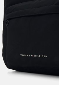 Černá nylonová taška s přední kapsou, jemnou texturou a tištěným logem "TOMMY HILFIGER" v bílé, červené a modré barvě.