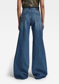 Džínové kalhoty s širokými nohavicemi v modré barvě, vysokým pasem, výrazným prošíváním na zadních kapsách a hladkou texturou, zobrazené ze zadní strany.