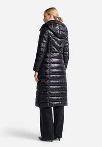 Sort pufferjakke med aftagelig hætte, kviltet design, sidelommer og lang længde, parret med brede sorte bukser og spidse hæle.