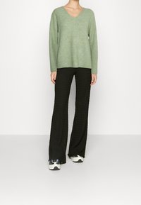 Pull vert clair à col en V avec manches longues, associé à un pantalon large noir texturé et des baskets blanches avec des accents noirs.