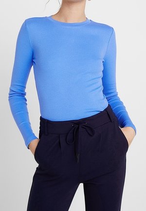 Kobieta ubrana w dopasowaną, długorękawową, prążkowaną niebieską bluzkę wsuniętą do wysokich, czarnych spodni z wiązanym paskiem w talii, z rękami w kieszeniach.