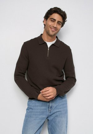 Hombre con cabello rizado oscuro sonríe llevando un suéter marrón oscuro con cremallera y mangas largas sobre una camisa blanca y vaqueros azul claro, frente a un fondo liso.