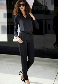 Zwarte blouse met lange mouwen en een kraag, gecombineerd met zwarte getailleerde broek. De broek heeft een trekkoord in de taille en subtiele zijzakken. Zwarte hakken.