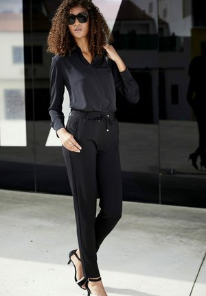 Schwarze Bluse mit langen Ärmeln und Kragen, kombiniert mit schmal zulaufenden schwarzen Hosen. Die Hosen verfügen über einen Kordelzugbund und dezenten Seitentaschen. Schwarze Absätze.