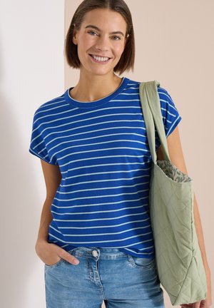 Femme souriante, portant un t-shirt rayé bleu, un jean bleu clair et portant un sac cabas matelassé vert sur l'épaule.