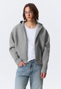 Harmaa vetoketjullinen huppari pehmeästä kankaasta, jossa rento istuvuus, suuret taskut ja nyörillä kiristettävä huppu, yhdistetty vaaleansinisiin farkkuihin.