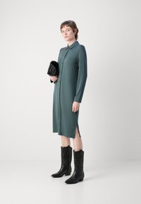 Robe longue à boutons en tissu vert foncé avec un col. Présente une fente sur le côté et est associée à des bottes de cowboy noires jusqu'au genou.