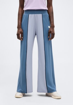 Άτομο που φοράει ψηλόμεσα παντελόνια με ψηλό ποδόγυρο και φαρδιά μπάρα, με κάθετες μπλε και ανοιχτογκρι λωρίδες, μαύρη περίμετρο, συνδυασμένα με λευκά αθλητικά παπούτσια.