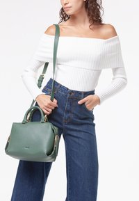 Borsa a tracolla in pelle verde con doppio manico e una tracolla, con hardware tono oro. Il modello indossa un top bianco a coste off-shoulder e jeans.
