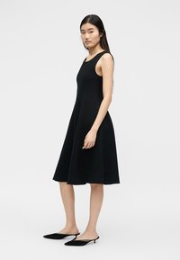 Γυναίκα στέκεται φορώντας μαύρο μίντι φόρεμα χωρίς μανίκια και μαύρα μυτερά mules με τακούνι, μπροστά σε απλό λευκό φόντο.