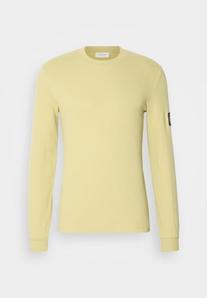 Tricou cu mânecă lungă de culoare galben deschis, fabricat dintr-un material texturat. Dispune de un gât rotund și un mic emblema pe mâneca stângă.