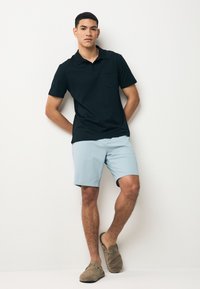 Musta poolopaita, jossa on tasku, yhdistetty vaaleansinisiin shortseihin. Mallilla päällä taupen väriset slip-on-kengät. Yksinkertainen muotoilu ja yhtenäiset värit.