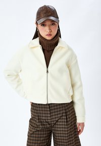 Krämfärgad zip-jacka med krage och ribbade nederkanter, kombinerad med en brun stickad polotröja och bruna rutiga byxor, under en brun keps.