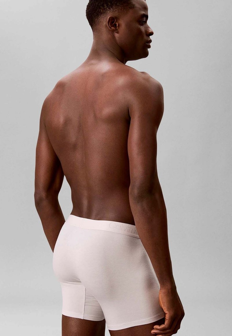 Atletische donkerharige man van achteren afgebeeld, gekleed in licht beige boxershorts tegen een effen grijze achtergrond.