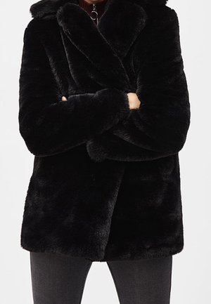 Personne portant un manteau en fausse fourrure noire avec un large col, bras croisés, associé à un pantalon gris foncé sur un fond uni.