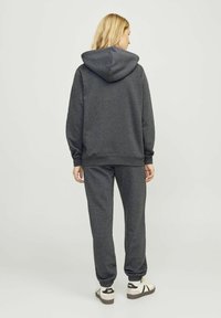 Mørk grå hettegenser og matchende joggebukser laget av myk stoff. Hettegenseren har en stor lomme, og buksene har elastiske mansjetter. Skoene er hvite med svarte detaljer.