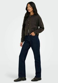 Dunkelbraune Wolljacke mit rundem Halsausschnitt, Knopfverschluss und gerippten Bündchen, kombiniert mit hoch geschnittenen dunkelblauen Jeans und schwarzen Stiefeletten.