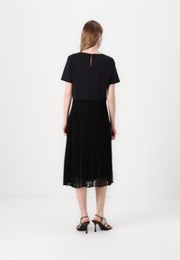 Abito nero con maniche corte; top strutturato con gonna plissettata; tessuto trasparente; dettaglio a chiave sulla schiena; indossato con tacchi alti neri.