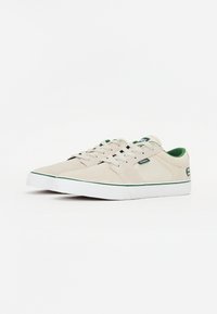Beige sneakers with textured upper, white rubber sole, green accents, and logo detail on the heel. Laced design with soft interior lining.