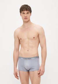 Pelēkas bokserbikses no gluda auduma ar ciešu pieguļumu, melnu jostas daļu ar baltā krāsā iespiestu uzrakstu "Calvin Klein".