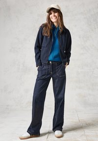 Námořnická zipová bunda, tyrkysový žebrovaný svetr, široké tmavě modré džíny, bílé tenisky s béžovými akcenty a světle zbarvená kšiltovka.