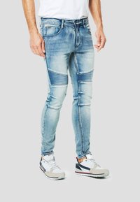 Dopasowane niebieskie dżinsy z denimu z prążkowanymi akcentami na kolanach, wyblakłym efektem i projektem z pięcioma kieszeniami. Noszone z białymi i szarymi butami sportowymi.