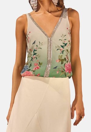 Haut sans manches à col en V avec motif floral vert et bordure métallique, associé à une jupe crème taille haute et de grandes boucles d'oreilles en forme de pétales.