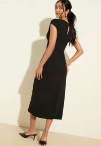 Schwarzes Midikleid mit kurzen Ärmeln, taillierter Oberteil, Rückenschlitz und seitlichen Schlitzen. Getragen mit schwarzen offenen Absatzschuhen.