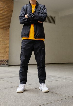 Person, der står med armene over kors iført en sort jakke og bukser, en gul trøje og hvide sneakers på et fliset gulv.
