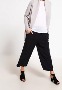 Öppen stickad cardigan i ljusgrått, matchad med lösa svarta culotter. Modellen bär svarta klacksandaler med utskärningar.