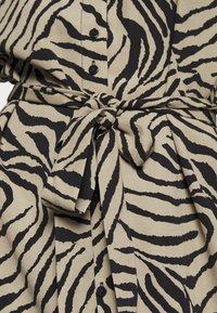 Písková tkanina s černým vzorem zebřích pruhů, s knoflíkovou přední částí a detailem vázání v pase. Textura vypadá hladce a lehce.