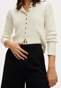 Top lungo a maniche ribbed color crema con scollatura bottonata e bottoni scuri a contrasto, abbinato a pantaloni neri sartoriali, che mostrano una vestibilità comoda.