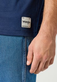 T-shirt Wrangler blu navy con scollo rotondo, maniche corte e un'etichetta in tessuto cucita sull'orlo. Indossato con jeans in denim azzurro chiaro.