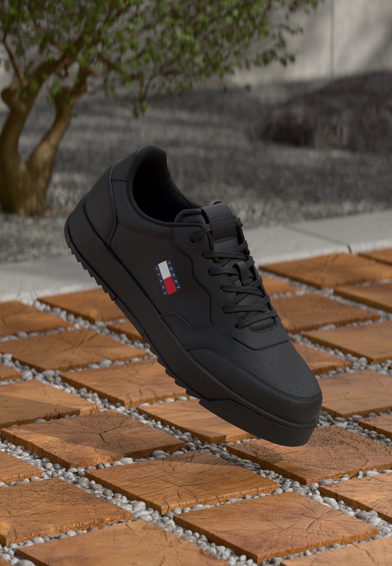 Baskets noires avec lacets et petit logo Tommy Hilfiger rouge, blanc et bleu flottant au-dessus de carreaux en bois en plein air.