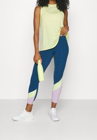 Světle zelený bezrukávový top, modré legíny se světle zelenými a levandulovými panely a bílé tenisky. Drží světle zelenou láhev na vodu.