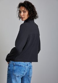 Musta lyhyt takki korkealla kauluksella ja pitkillä hihoilla, yhdistettynä vaaleansinisiin farkkuihin, joissa on takataskut ja kulunut pinta.