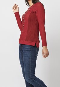 Haut rouge à manches longues avec un design texturé, un laçage au niveau de l'encolure et des fentes sur les côtés. Associé à un jean bleu foncé ajusté.