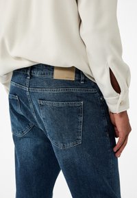 Dżinsy w ciemnoniebieskim kolorze o gładkiej fakturze, z tylnymi kieszeniami i beżową skórzaną łatą na pasku. Model ma na sobie jasny top.