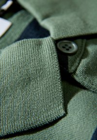 Zielony dziany kardigan z fakturowanym kołnierzem, z szarym detalem z guzików i gładką tkaniną. Widok z bliska podkreśla wzory szwów.