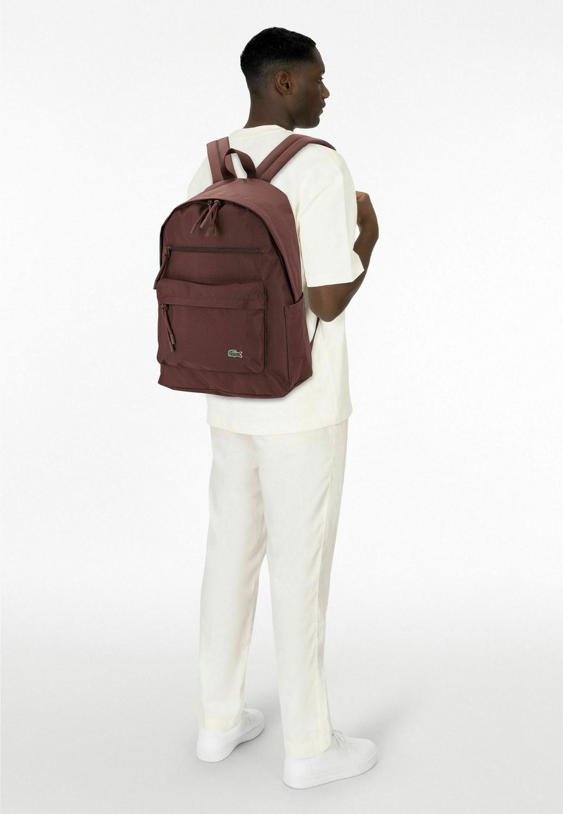 Maroon backpack with textured fabric, front zipper pocket, two shoulder straps, and a small logo detail. Worn by a person in a white outfit.