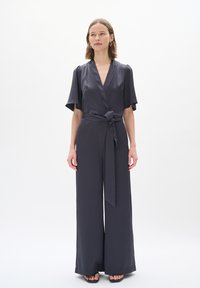 Jumpsuit avvolgente grigio scuro con maniche corte, vita con fiocco, gambe larghe e una finitura testurizzata. Ambientato su uno sfondo bianco semplice.