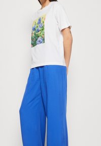 Bílá bavlněná trička s barevným květinovým potiskem. Spárované s modrými širokými kalhotami. Jednoduchý design s krátkými rukávy a kulatým výstřihem.