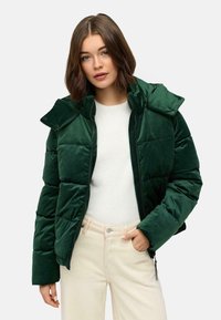 Groene fluwelen pufferjack met hoge kraag, ritssluiting en drukknoopjes. Gepaard met een lichtgekleurde trui en beige broek.