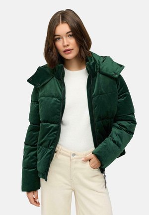 Groene fluwelen pufferjack met hoge kraag, ritssluiting en drukknoopjes. Gepaard met een lichtgekleurde trui en beige broek.