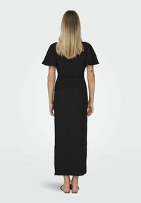 Vestido maxi de punto negro con mangas cortas acampanadas, cuerpo ajustado, tela texturizada y una silueta recta, llevado sobre sandalias planas.