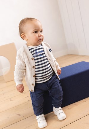 Cardigan lavorato a maglia beige chiaro con bottoni, camicia a righe navy e bianca, pantaloni cargo navy e scarpe bianche su uno sfondo semplice.